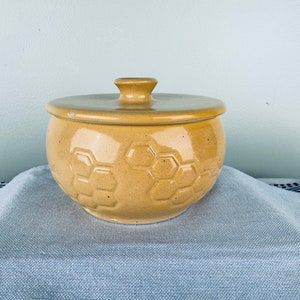 May include: A yellow ceramic honey pot with a lid. The pot has a honeycomb pattern.