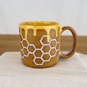 May include: A ceramic mug with a honey-themed design. The mug is brown with a textured surface and features a honeycomb pattern outlined in white. The rim and drips of honey are a golden yellow color. The handle is brown.