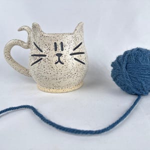 May include: A speckled ceramic cat mug with a handle and cat face design, next to a ball of blue yarn. The mug has a cream-colored base with black speckles and black facial features. The yarn is a deep blue color.