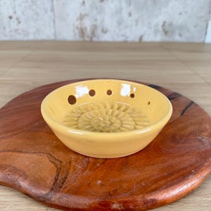 May include: A small, round, yellow ceramic bowl featuring a series of small circular holes along the upper rim and a raised, sunburst-like floral pattern embossed on the interior base. The bowl has a smooth, glossy finish and a shallow, open design.