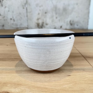 May include: A white ceramic bowl with a speckled finish and a dark brown base. Black chopsticks rest across the top of the bowl. The bowl has a unique design with a notch to hold the chopsticks.