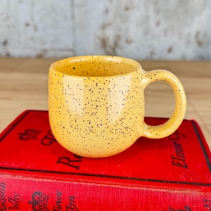 May include: A yellow speckled ceramic mug with a rounded body and a handle. The mug is sitting on top of a red book with gold lettering. The mug has a capacity of approximately 10 ounces.