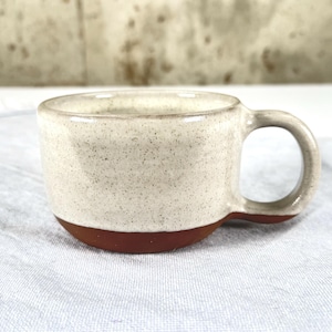 May include: A handmade ceramic mug with a speckled, off-white exterior and a reddish-brown base. The mug has a rounded body, a sturdy handle, and is designed for hot beverages. The mug is approximately 3 inches tall.