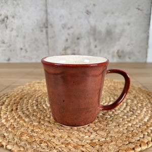 May include: A ceramic mug with a glossy, deep reddish-brown exterior glaze and a contrasting speckled off-white interior. The mug features a slightly flared rim, a rounded handle on the right side, and a smooth, tapered body with subtle vertical indentations.