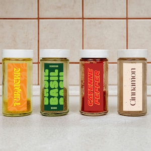 May include: Four glass spice jars with white lids are displayed. The labels read "Turmeric", "Basil", "Cayenne Pepper", and "Cinnamon". The jars are arranged on a white surface against a backdrop of beige square tiles.