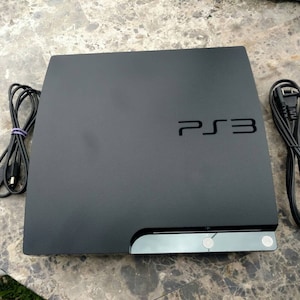 May include: A black PS3 console with the text "PS3" on the top. The console has a sleek design with a silver trim and two cables, one with a USB connector and the other with a power plug.