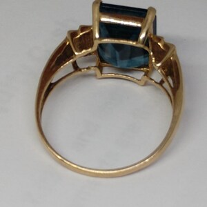 May include: A gold ring with a large, dark blue, emerald-cut gemstone. The ring has a simple band and a delicate, ornate setting for the stone.