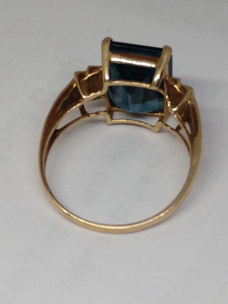 May include: A gold ring with a large, dark blue, emerald-cut gemstone. The ring has a simple band and a delicate, ornate setting for the stone.