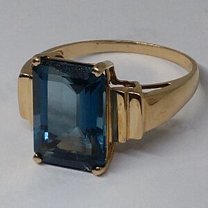 May include: A gold ring with a large, rectangular, blue gemstone. The ring has a simple band with a slightly raised edge.