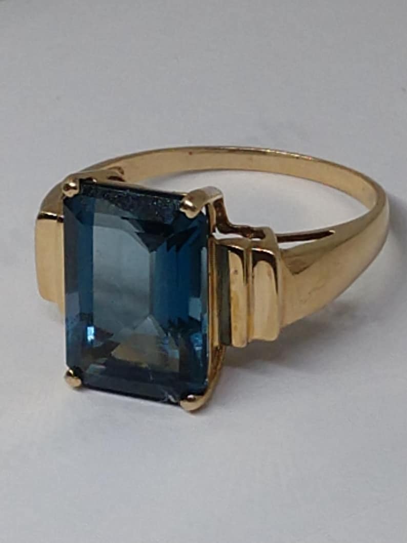 May include: A gold ring with a large, rectangular, blue gemstone. The ring has a simple band with a slightly raised edge.