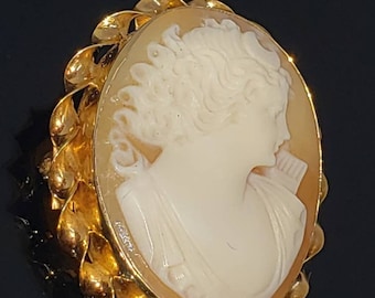 May include: A gold cameo pendant featuring a carved portrait of a woman with curly hair. The cameo is set in a gold frame with a decorative leaf design.