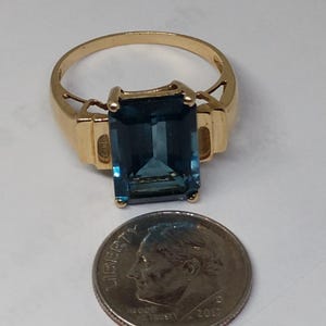 May include: A gold ring with a large, dark blue, emerald-cut gemstone. The ring is shown next to a US dime for size comparison.