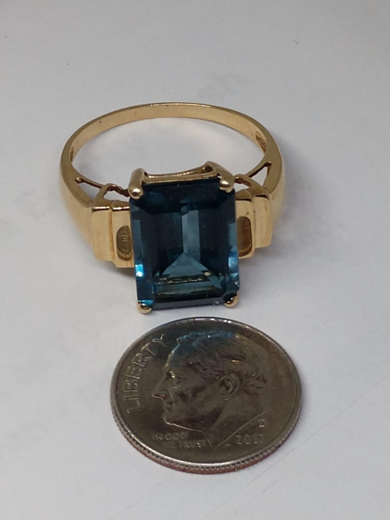 May include: A gold ring with a large, dark blue, emerald-cut gemstone. The ring is shown next to a US dime for size comparison.