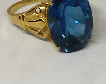 May include: A gold ring with a large, oval-shaped blue gemstone. The ring has a detailed, ornate design.