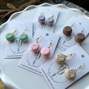 May include: Assortment of handmade polymer clay earrings shaped like macarons. The earrings are in various colors, including pink, green, brown, and beige. Some macarons have sprinkles. The earrings are displayed on white cards with the text "Jewelry and Accessories" and "mydesiertodesigns.etsy.com".