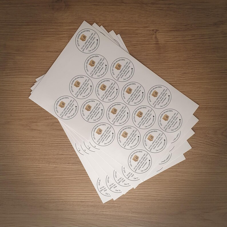 Puede incluir: Una pila de hojas de papel blancas con pegatinas redondas marrones. Cada pegatina tiene una caja marr&oacute;n con un coraz&oacute;n y el texto "Ich bin wiederverwendbar. Der Umwelt zuliebe."