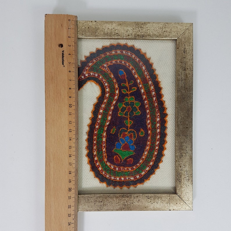 May include: A framed artwork featuring a colorful paisley design with intricate details. The paisley is embroidered on a white fabric and framed in a silver-colored frame.