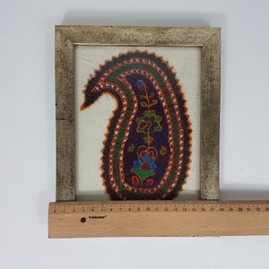 May include: A framed artwork featuring a colorful paisley design embroidered on a white fabric. The paisley is outlined in orange and green, with a floral motif in the center.