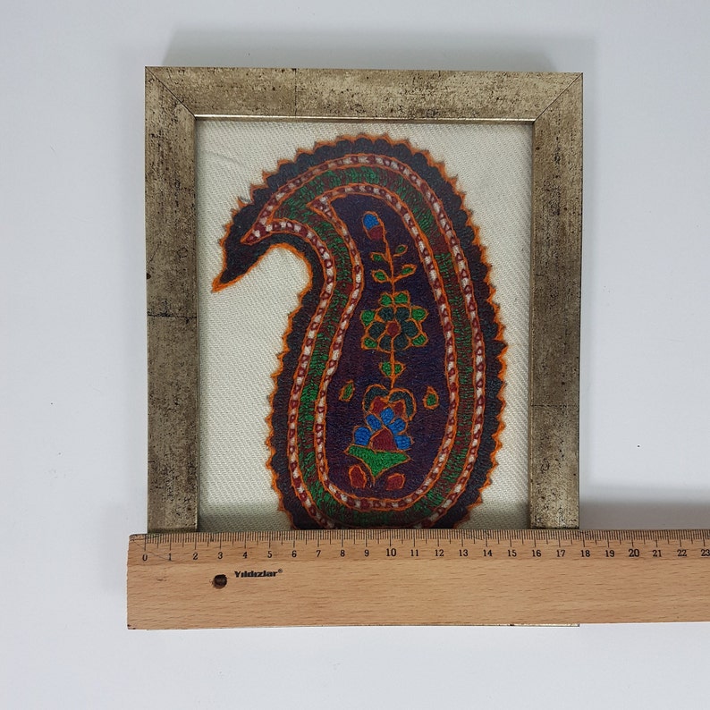 May include: A framed artwork featuring a colorful paisley design embroidered on a white fabric. The paisley is outlined in orange and green, with a floral motif in the center.
