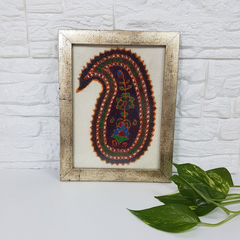 May include: A framed artwork featuring a colorful paisley design with intricate details. The paisley is embroidered on a white fabric and framed in a silver-colored frame.