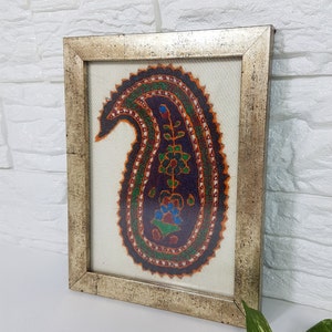 May include: A framed embroidery featuring a colorful paisley design in shades of orange, green, blue, and purple. The paisley is surrounded by a white background and a silver frame.