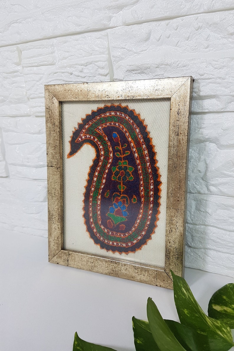 May include: A framed embroidery featuring a colorful paisley design in shades of orange, green, blue, and purple. The paisley is surrounded by a white background and a silver frame.