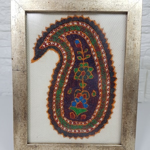 May include: A framed embroidery of a paisley design in shades of orange, green, blue, and red on a white background. The frame is silver with a gold trim.