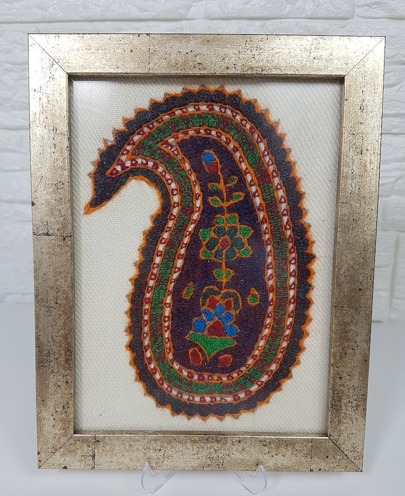 May include: A framed embroidery of a paisley design in shades of orange, green, blue, and red on a white background. The frame is silver with a gold trim.