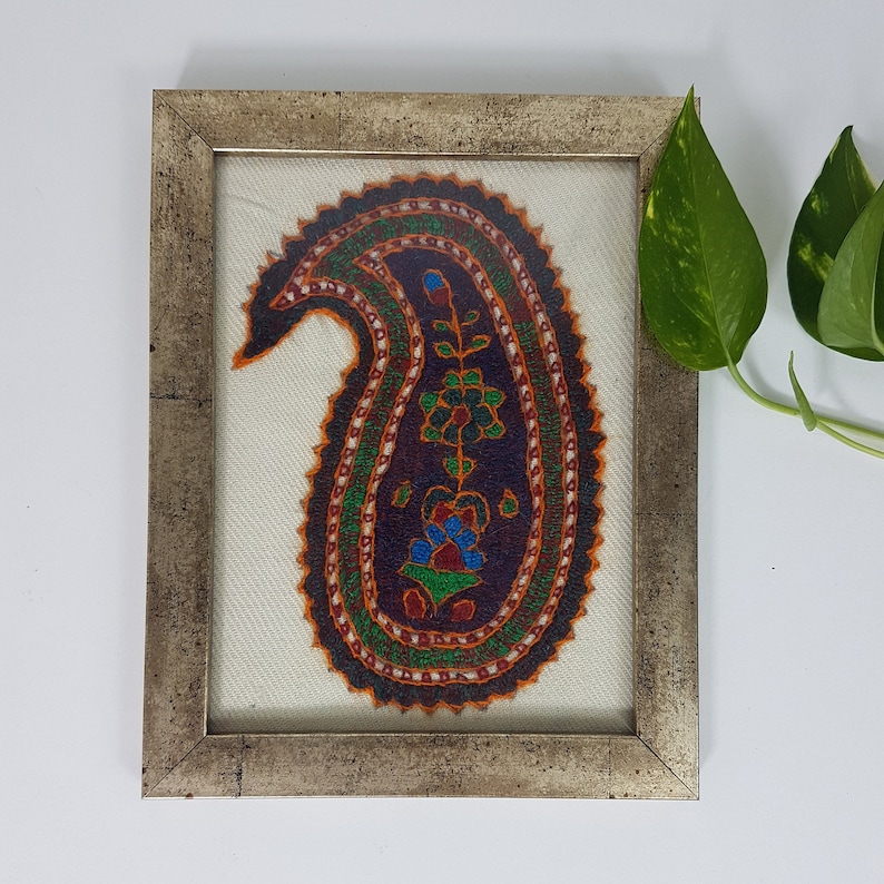 May include: A framed artwork featuring a colorful paisley design embroidered on a white fabric background. The paisley is outlined in black and filled with intricate details in shades of orange, green, blue, and red.