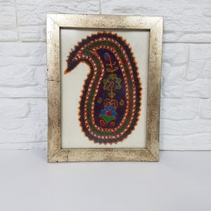 May include: A framed artwork featuring a colorful paisley design with intricate details. The paisley is outlined in orange and green, with a dark purple center and a floral motif in the center. The artwork is framed in a silver frame.