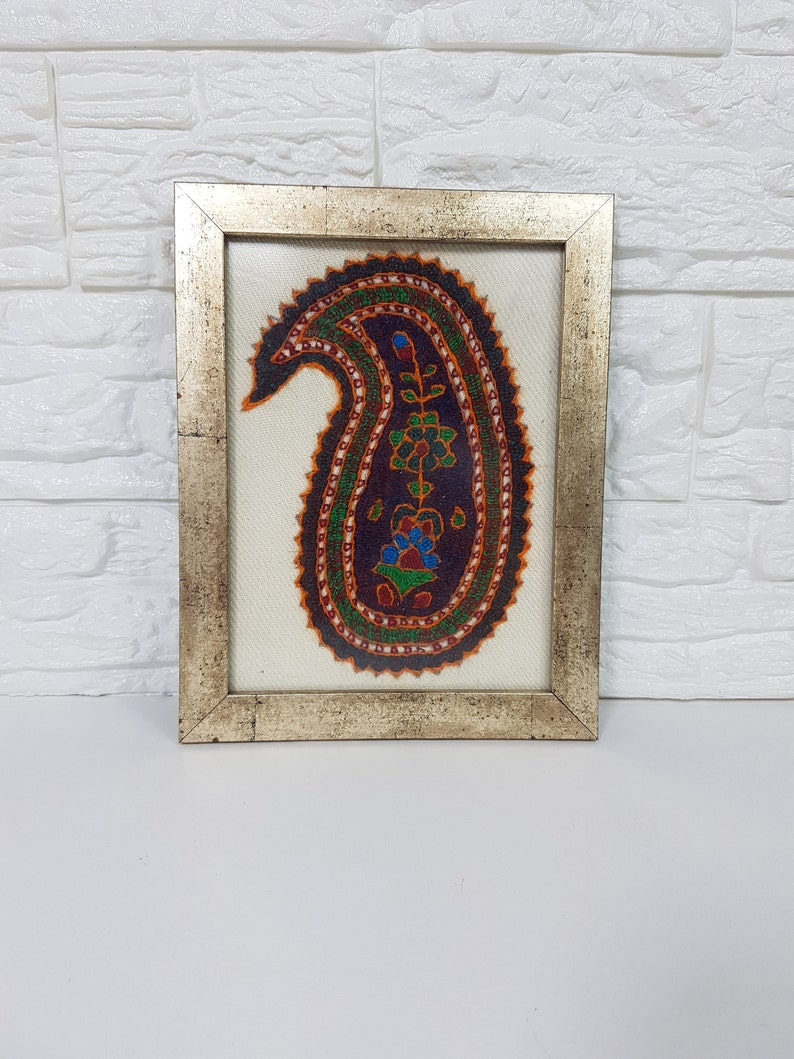 May include: A framed artwork featuring a colorful paisley design with intricate details. The paisley is outlined in orange and green, with a dark purple center and a floral motif in the center. The artwork is framed in a silver frame.