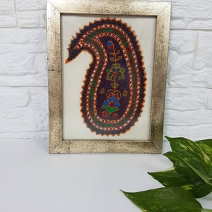 May include: A framed artwork featuring a colorful paisley design embroidered on a white background. The paisley is outlined in black and filled with shades of red, orange, green, blue, and purple.