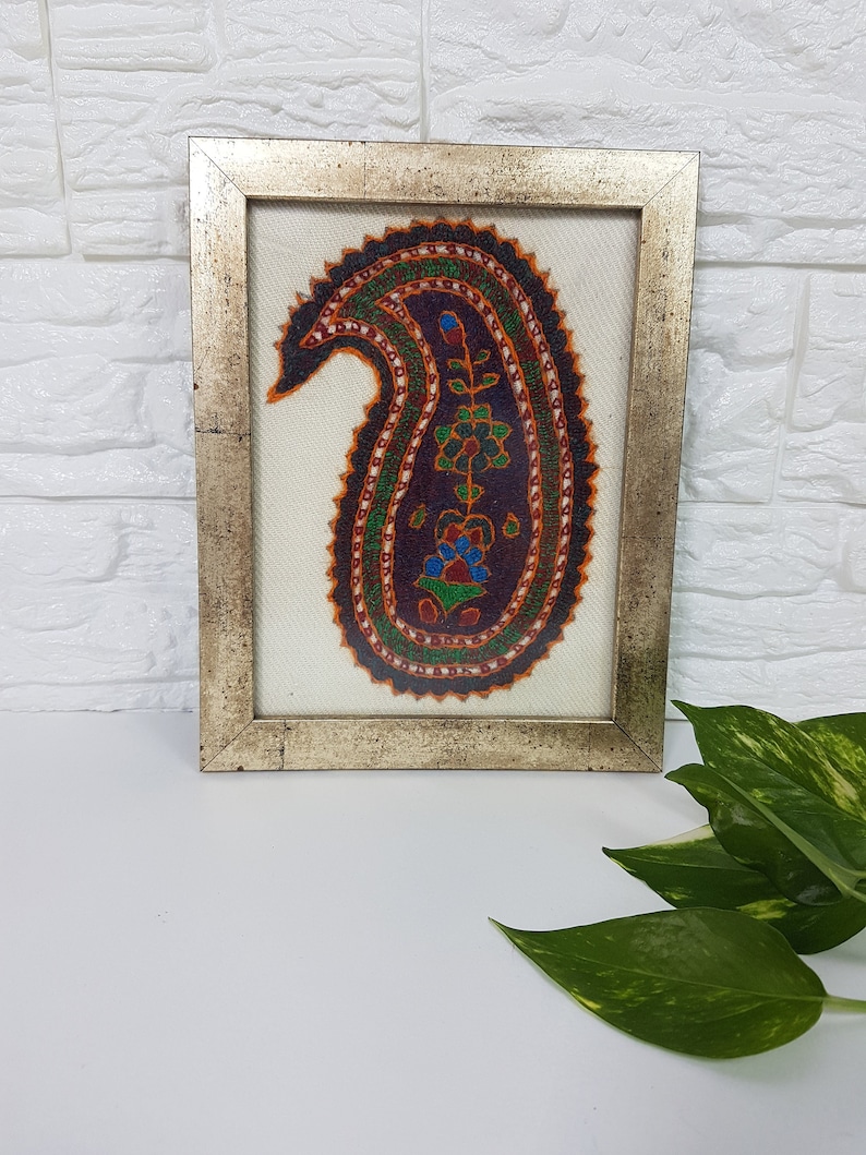 May include: A framed artwork featuring a colorful paisley design embroidered on a white background. The paisley is outlined in black and filled with shades of red, orange, green, blue, and purple.