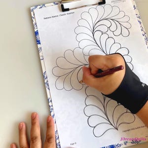 May include: A clipboard with a floral design border holds a printed pattern titled "Classic Feather." A hand wearing a black wrist brace is drawing on the pattern with a pen. The pattern features a feather design with dotted lines.