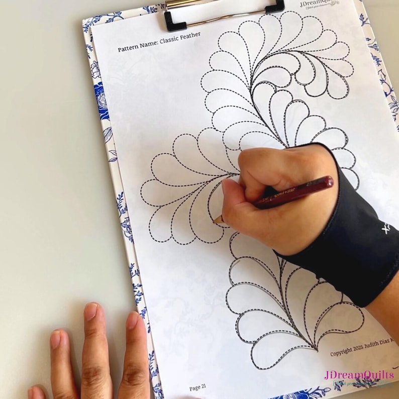 May include: A clipboard with a floral design border holds a printed pattern titled "Classic Feather." A hand wearing a black wrist brace is drawing on the pattern with a pen. The pattern features a feather design with dotted lines.