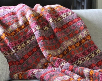May include: A colorful knitted blanket draped over a sofa. The blanket features horizontal stripes in shades of pink, orange, and gray. A red vase with flowers and a decorative stone pot are visible in the background.