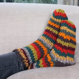 May include: A pair of colorful striped knitted socks in shades of red, orange, yellow, green, brown, and gray. The socks are resting on a gray couch with a herringbone pattern.