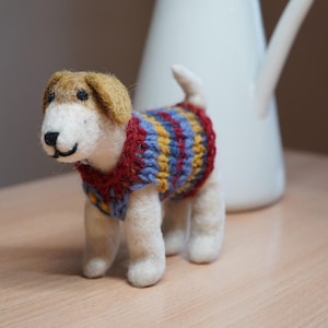 Könnte beinhalten: Filzhunde-Figur mit farbenfrohem Pullover. Der Hund hat einen weißen Körper, braune Ohren und eine schwarze Nase. Der Pullover ist rot, blau und gelb. Eine weiße Kanne mit lila Blumen befindet sich im Hintergrund.