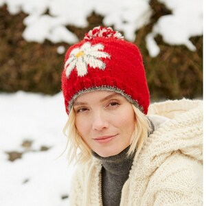 May include: A red knit beanie with a white daisy design and a white pom pom on top. The beanie is worn by a person with blonde hair and a white cable knit sweater.