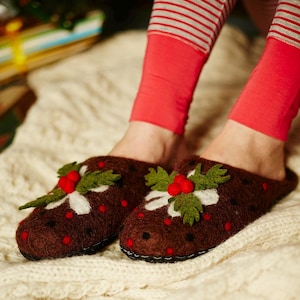 May include: A pair of brown felt slippers with a Christmas pudding design. The slippers have green holly leaves, red berries, and white icing details.