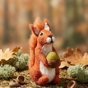 Peut inclure: Une figurine d'écureuil en laine feutrée à l'aiguille, composée de fibres orange et blanches. L'écureuil est assis et tient un petit gland feutré vert et marron entre ses pattes avant. Il possède des yeux en perles noires et une queue touffue.
