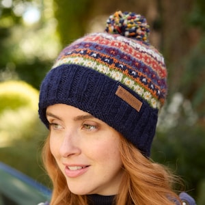 Könnte beinhalten: Eine marineblaue Strickmütze mit bunt gemustertem Oberteil und Bommel. Die Mütze hat einen braunen Lederaufnäher mit dem Markennamen. Die Mütze ist für kaltes Wetter konzipiert.
