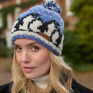 Op de afbeelding: Een blauwe en witte gebreide muts met een pompon bovenop. De muts heeft een patroon van pinguïns en sneeuwvlokken. De muts heeft een bruin lederen label met de tekst "ROCHEMAN" erop.