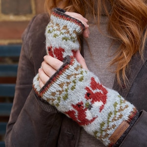 May include: A pair of grey and white knitted fingerless gloves with a red fox design. The gloves have a brown leather tag with the text "The Handmade Shop" on it.