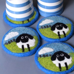 Puede incluir: Cuatro posavasos de cerámica a rayas azules y blancas con un diseño de oveja de fieltro. Cada posavasos presenta una oveja blanca y negra de pie en un campo verde con un cielo azul y nubes blancas.