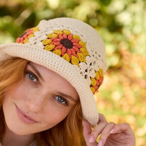 May include: A crocheted hat with a wide brim and a floral design in shades of orange, yellow, and brown. The hat is made with a white yarn and has a crocheted flower pattern around the brim.