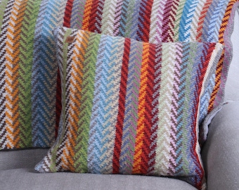 Könnte beinhalten: Zwei bunte Kissen mit Chevron-Muster auf einem grauen Sofa. Die Kissen sind aus einem weichen, gewebten Stoff gefertigt und in verschiedenen Farben gehalten, darunter Rot, Orange, Gelb, Grün, Blau und Lila.