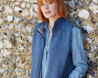 May include: A blue knitted vest with a zipper closure. The vest has a stand-up collar and is worn over a blue denim shirt. The person is standing in front of a stone wall.