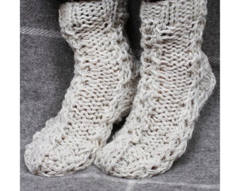 May include: A pair of cream-colored knitted socks with a cable knit pattern. The socks are worn on a person's feet and are resting on a gray and white patterned surface.