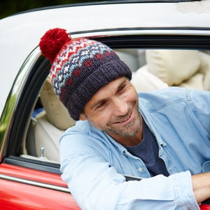 Può includere: Un uomo con una camicia blu e un berretto di lana rosso, bianco e blu con un pon pon in cima, sorride mentre guarda fuori dal finestrino di un'auto rossa.